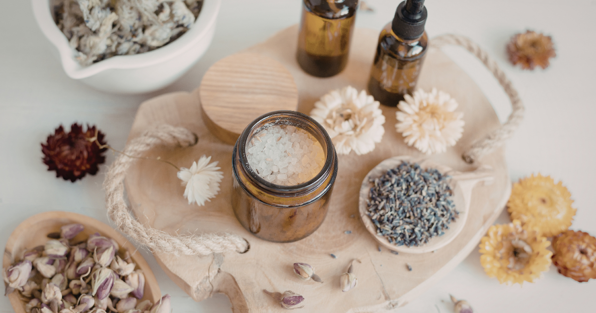 Ästhetisches Arrangement für DIY Kosmetik mit Badezusatz, ätherischen Ölen in Braunglasflaschen und Trockenblumen auf einem Holztablett.